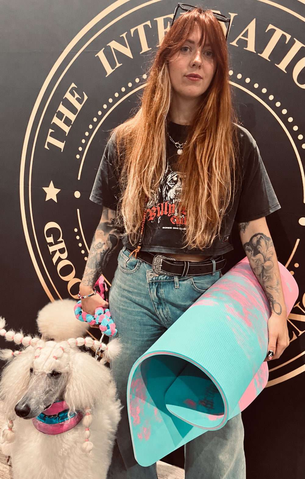 Groomer holding the rolled reversible green and pink anti-fatigue mat beside a white poodle